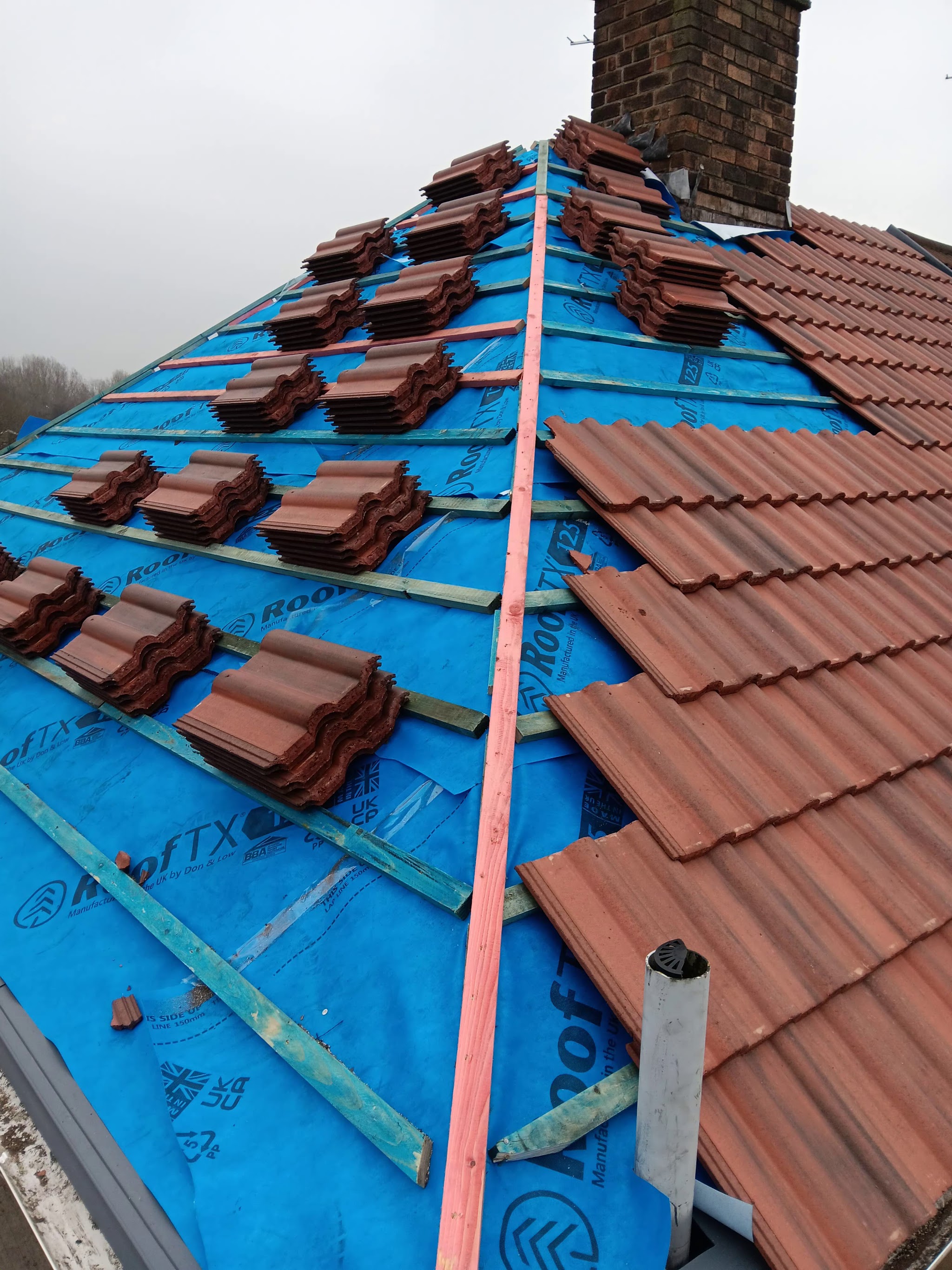 Red interlocking roof tiles stacked on battens and being laid beside a chimney